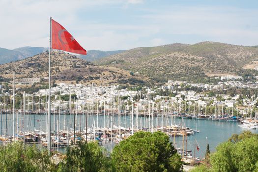 View of Bodrum harbour and marina from St Peter's castle, in Mugla Province, Turkey. The Turkish flag is on the foreground and the ruins of an ancient theatre can be seen in the background.