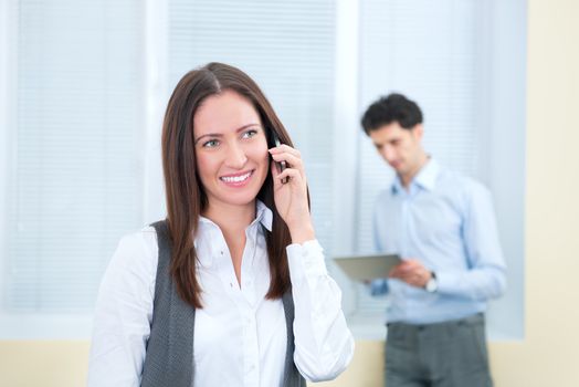 Beautiful young business woman speaks on a mobile phone. Young businessman with digital tablet on the background in soft focus.
