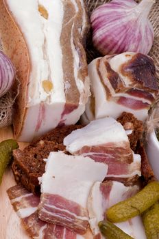 Arrangement of Sliced Raw Smoked Homemade Bacon, Garlic, Brown Bread and Marinated Gherkins closeup on Cutting Board