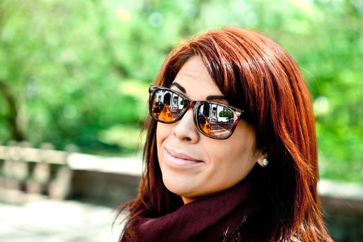 Young woman standing in front of green park trees with bright red hair headshot portrait. Photographed in the city with reflections in the lenses showing urban life.