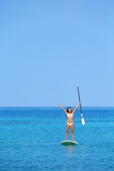 Aspirational beach lifestyle woman on paddleboard enjoying summer holidays vacation in bikini on hawaii. Beautiful mixed race girl on hawaiian paddleboard with arms raised in happiness.