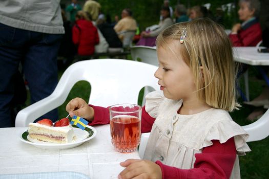 A little girl eating a strawberry cake decorated with the swedish flat. She is celebrating midsummer. She also has a glass of strawberry squash