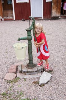 A cute blonde little girl standing next to a waterpump at an outdoor museum
