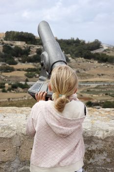 A girl standing at a viewpoint in Mdina, Malta, with her back to the camera, looking through a viewpoint telescope at the view