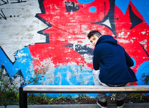 Young man with hoodie sitting on bench in front of graffiti wall