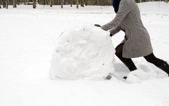 woman in grey coat push big snow ball roll in winter meadow. snowman building.