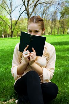 Woman hides her face behind a book sitting on green grass