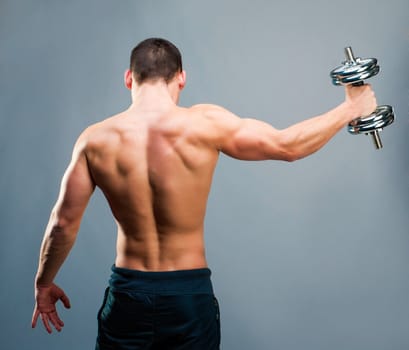 Rear view of a young male bodybuilder doing heavy weight exercise with dumbbells against gray backg