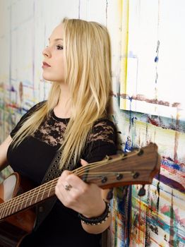 Photo of a young blond female holding an acoustic guitar and leaning up against a wall.