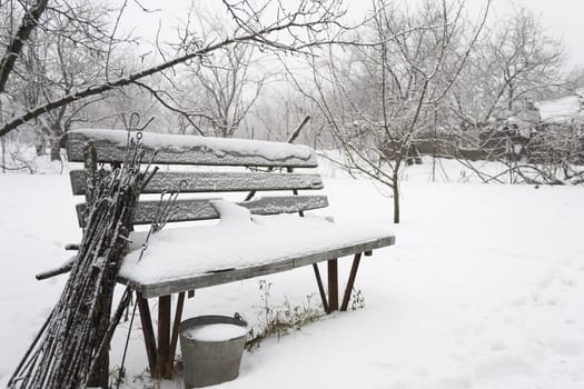 Snow-covered bench with forgotten garden tools in the winter garden.