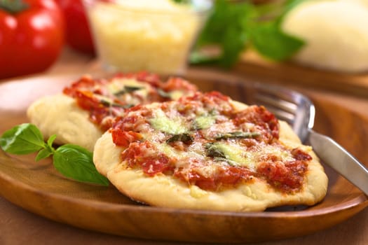 Crispy homemade Pizza Margherita or Pizza Mozzarella (pizza with tomato, basil and cheese) with fork on wooden plate with ingredients in the back (Selective Focus, Focus one third into the first pizza)