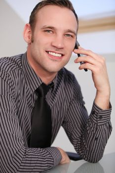 Close-up of a young business man smiling at the camera and talking on a smartphone. Smiling young man in a stylish shirt and tie sitting at a table using a smartphone or mobile phone