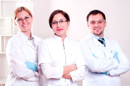 Three doctors are smiling at the camera in a doctors' office. Horizontally framed shot.