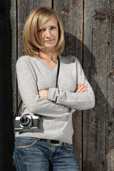 Photo of a blond female leaning up against an old barn with a antique camera on her shoulder.