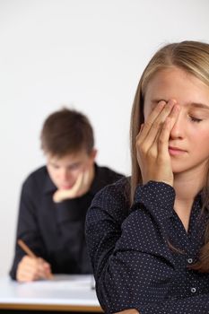 Photo of a very tired female student in class with a male student writing at a desk behind her.