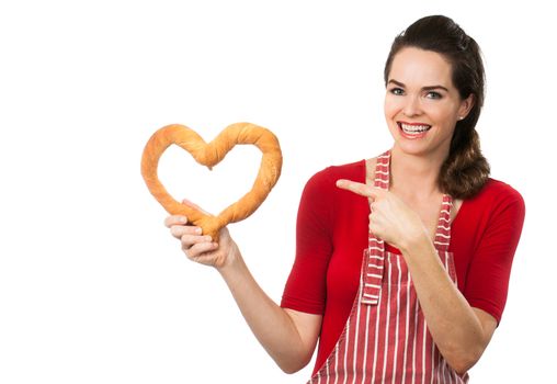 A beautiful woman wearing an apron smiling and pointing  at a love heart made out of bread. Isolated on white.