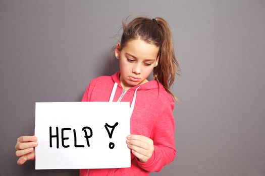 Conceptual image of a sad dejected little girl with a pouting lip standing holding a handwritten HELP sign with an exclamation mark, studio upper body portrait on a grey background with copyspace