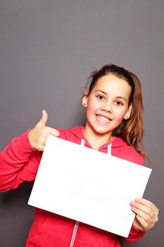 Beautiful smiling happy little girl with blank white sign giving a thumbs up of approval and agreement, studio upper body portrait on a grey background with copyspace