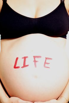 LIFE written on a womans bare pregnant belly in red lettering signifying the new life of her unborn child as its heart beats inside her, closeup cropped image of her tummy