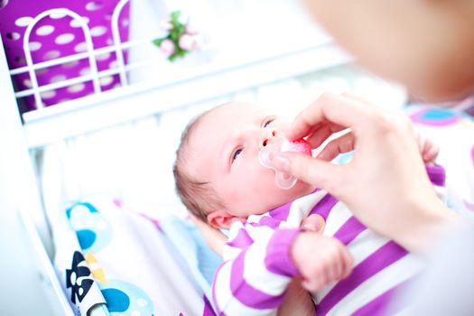 Loving young mother leaning over into a cot putting a dummy or pacifier into a beautiful newborn baby's mouth