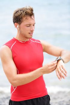 Running man looking at heart rate monitor GPS watch. Runner on beach listening to music in earphones wearing red compression t-shirt top.