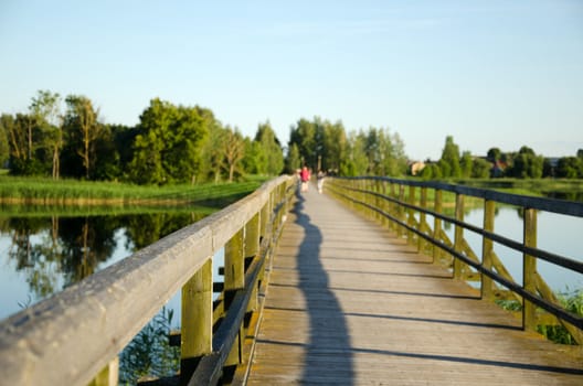 long wooden footbridge with railings over lake and blurred people walking in distance.