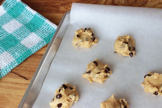 A close-up image of a cookie sheet lined with parchment paper, with chocolate chip cookie dough shaped into cookies, on a wooden counter with a green and white dish towel.