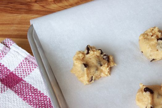 A close-up image of a cookie sheet lined with parchment paper, with chocolate chip cookie dough shaped into cookies, on a wooden counter with a red and white dish towel.