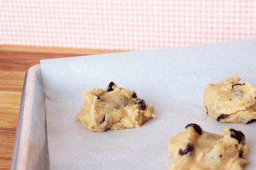 A close-up image of a cookie sheet lined with parchment paper, with chocolate chip cookie dough shaped into cookies, on a wooden counter with a pink and white gingham wallpaper background.