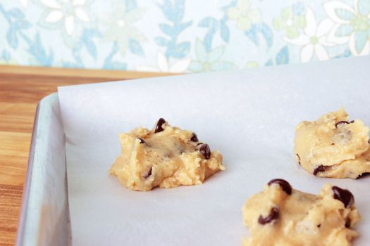 A close-up image of a cookie sheet lined with parchment paper, with chocolate chip cookie dough shaped into cookies, on a wooden counter with a vintage blue, white, and green flowered wallpaper background.