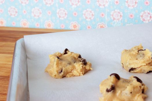 A close-up image of a cookie sheet lined with parchment paper, with chocolate chip cookie dough shaped into cookies, on a wooden counter with a vintage blue and pink flowered wallpaper background.