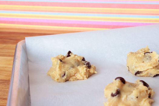 A close-up image of a cookie sheet lined with parchment paper, with chocolate chip cookie dough shaped into cookies, on a wooden counter with a pink and orange striped wallpaper background.