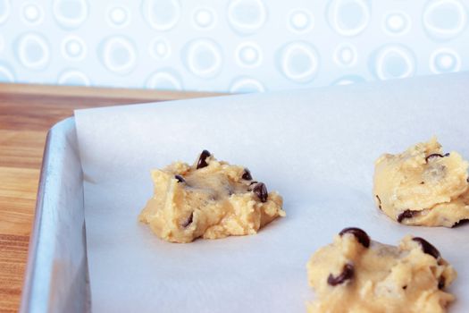 A close-up image of a cookie sheet lined with parchment paper, with chocolate chip cookie dough shaped into cookies, on a wooden counter with a modern blue and white wallpaper background.