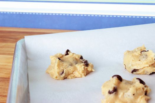 A close-up image of a cookie sheet lined with parchment paper, with chocolate chip cookie dough shaped into cookies, on a wooden counter with a modern blue striped wallpaper background.