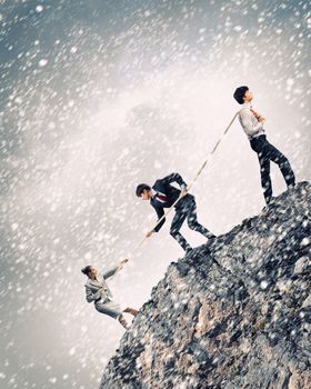 Image of three businesspeople pulling rope atop of mountain under falling snow