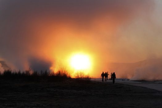 Silhouetted figures walking through steam from geysers with an orange glowing sunset