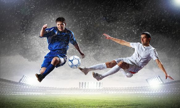 two football players in jump to strike the ball at the stadium under rain