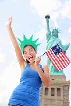 Tourist at Statue of Liberty, New York, USA standing with american flag excited and happy. Tourism, travel or patriotic concept with joyful multiracial woman.