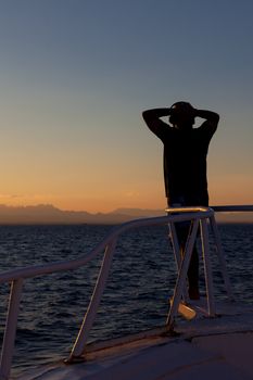 Person enjoying an ocean sunset standing on the prow of a ship looking towards the distant land and setting sun