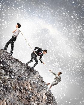 Image of three businesspeople pulling rope atop of mountain under falling snow