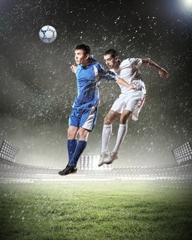 two football players in jump to strike the ball at the stadium under rain