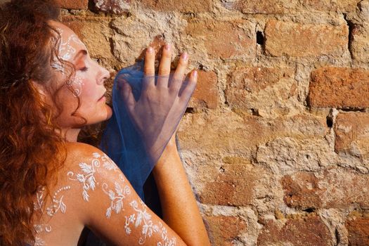Topless redhead woman with white body henna tattoo leaning against brick wall, eyes closed