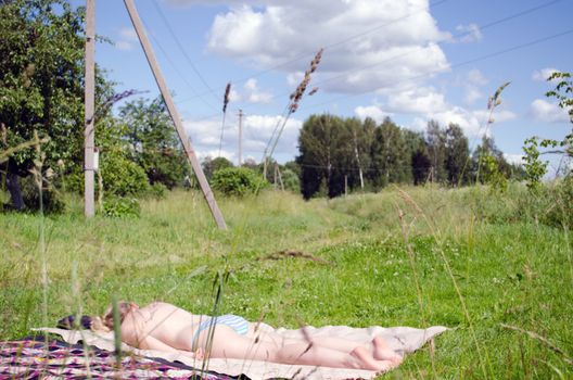 Half nude woman without bra visible through grass sunbathing on plaid in rural meadow.