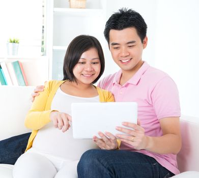 Asian couple. Husband and 8 months pregnant wife using tablet computer sitting on sofa at home