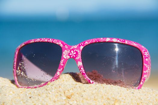 pink kids suglasses with a white floral design on the sand near the sea