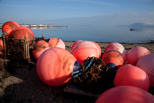 A group of orange mooring buoys arranged on the shore with a beach and the sea in the distance.