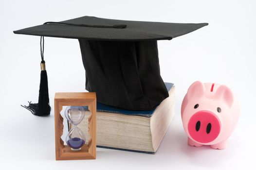 Graduation cap on a thick book with hourglass and piggy  bank