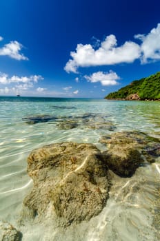 Rocks in clear Caribbean water with a yacht and lush green tropical island in the background