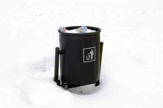 full garbage dustbin waste bin surrounded by snow in winter park.