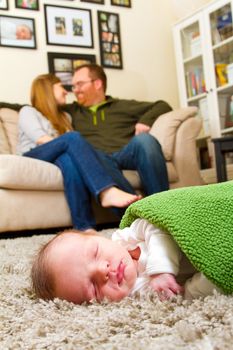A newborn baby boy sleeps on a comfortable rug while wrapped up in a green blanket with his parents in the background together.